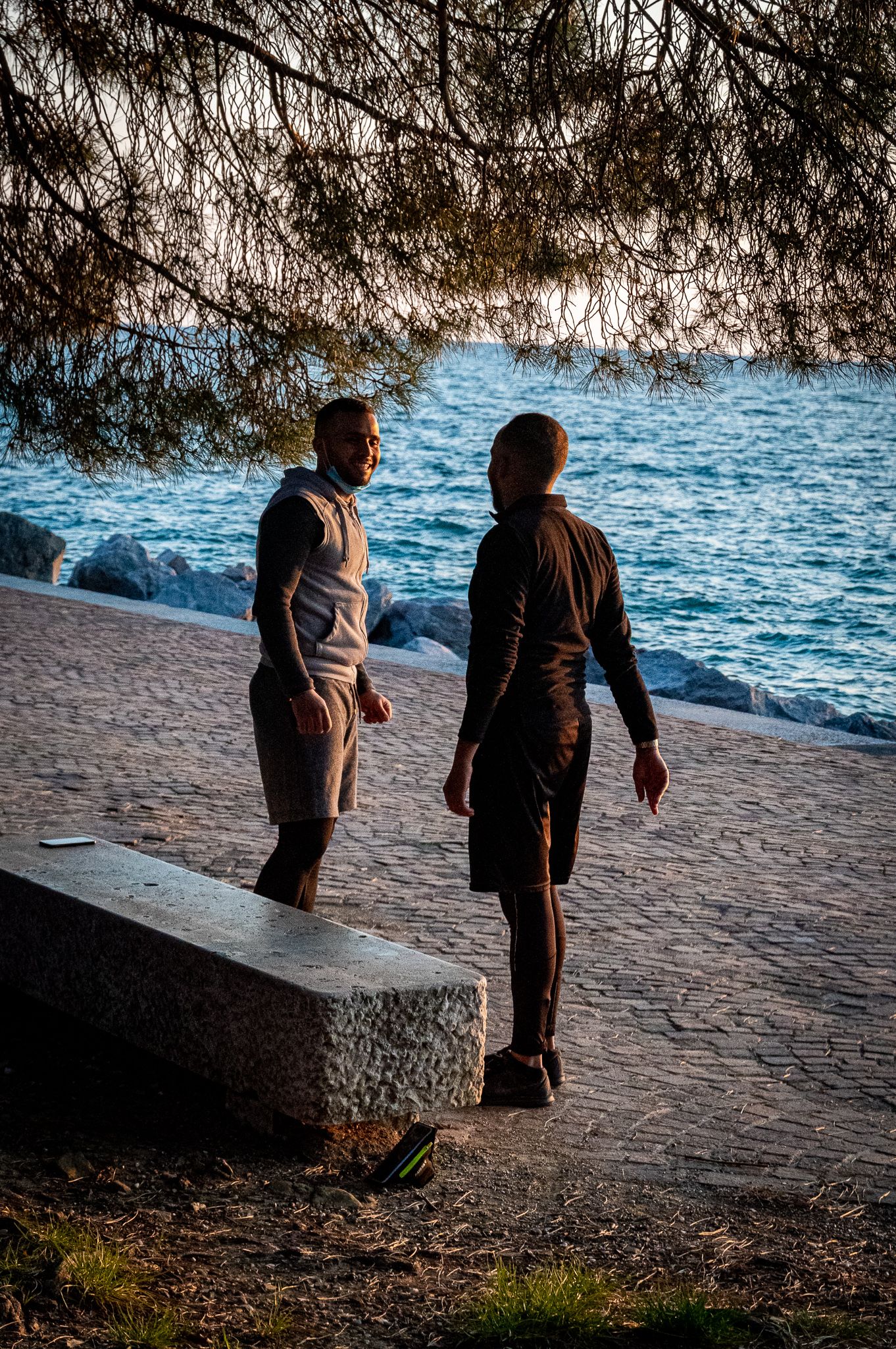 Two men standing on cobblestone waterfront promenade near rocky shoreline at sunset.