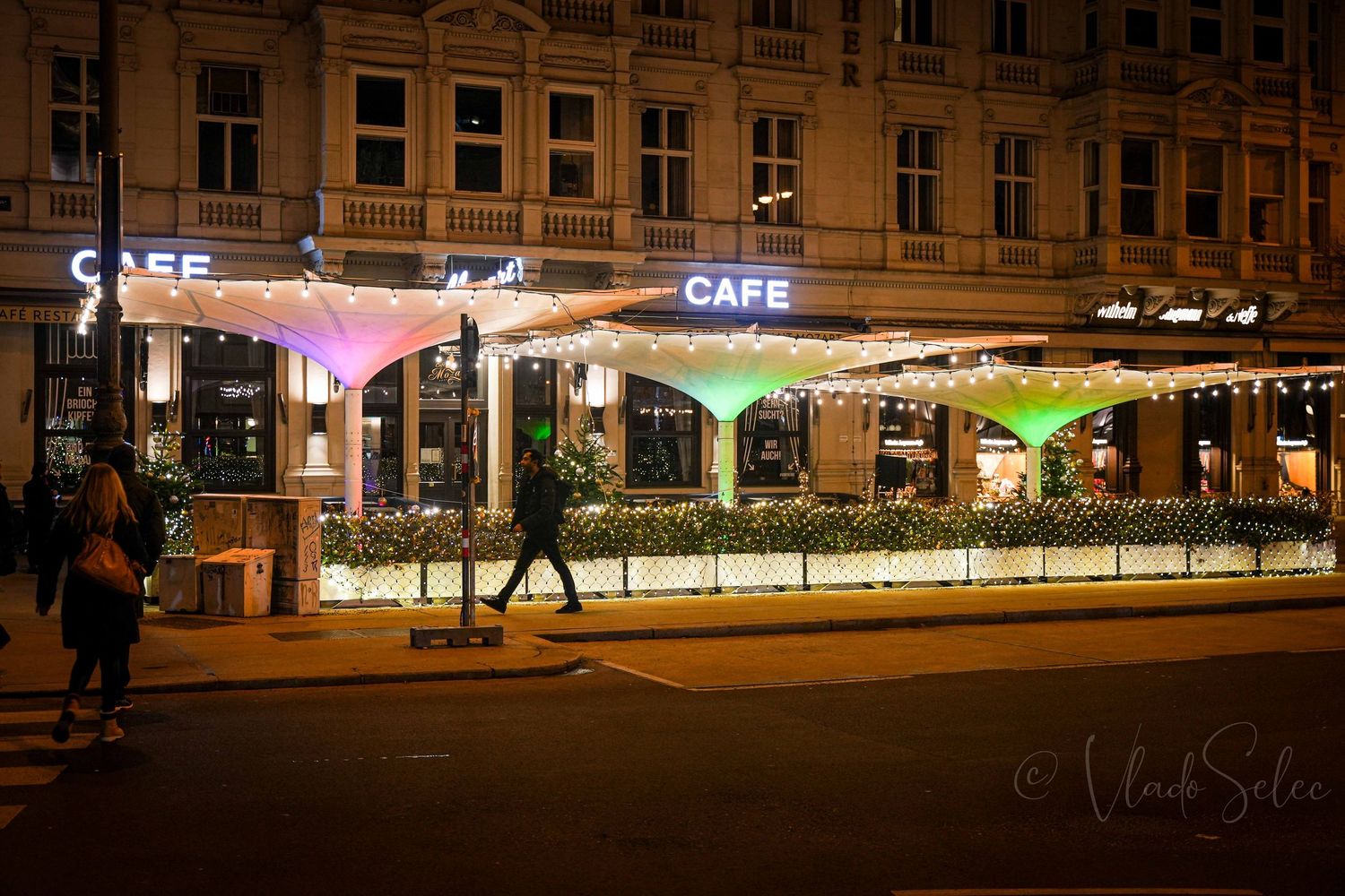 Colorful illuminated outdoor cafe terrace at night with string lights and vibrant green and pink canopies on a city street.