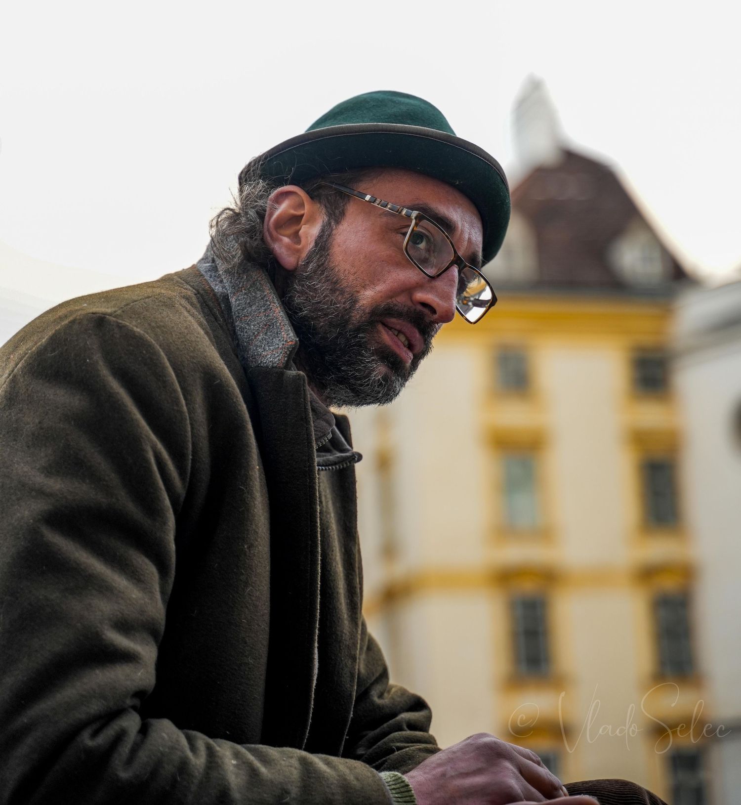 Bearded man wearing green hat and glasses in thoughtful pose outdoors in European city.