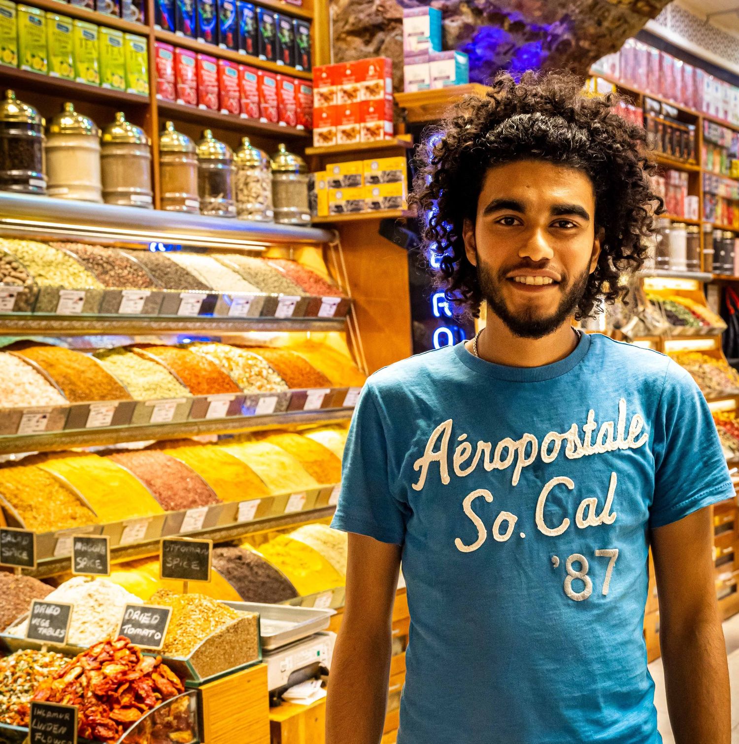 Smiling young man in Aeropostale t-shirt standing in front of colorful spice market display.