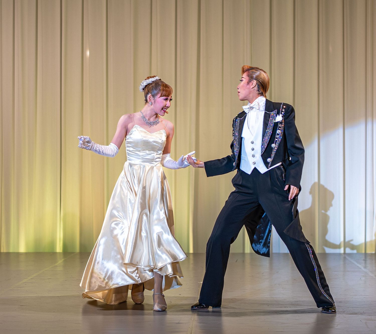 Two performers dancing on stage in formal attire, woman in white gown and man in black suit.