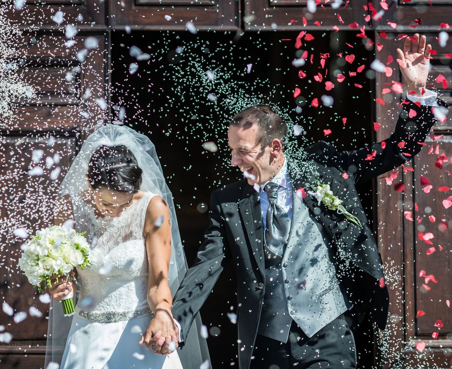 Joyful newlyweds exit church amid colorful confetti shower, bride holding white bouquet in elegant wedding attire.