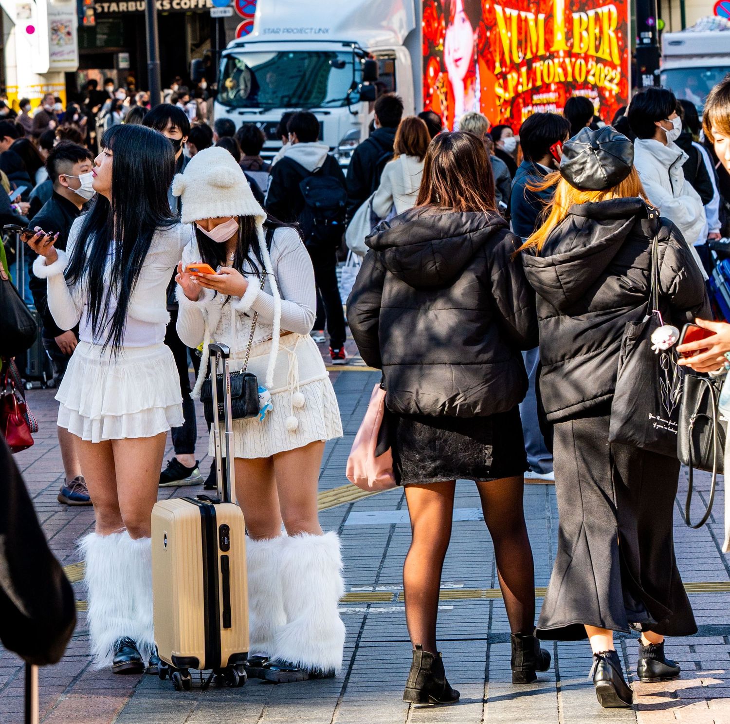 Stylish young women in white outfits with fur boots and luggage at a busy Tokyo street crossing.