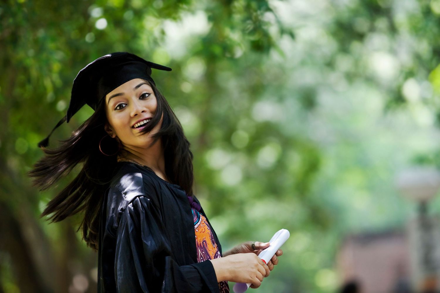 Smiling young Black woman in graduation cap and gown holding a diploma outdoors.