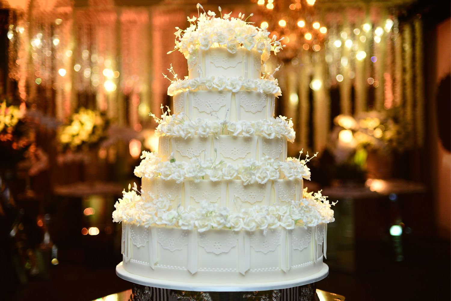 Elegant five-tier white wedding cake decorated with flowers and sparkling lights at reception.