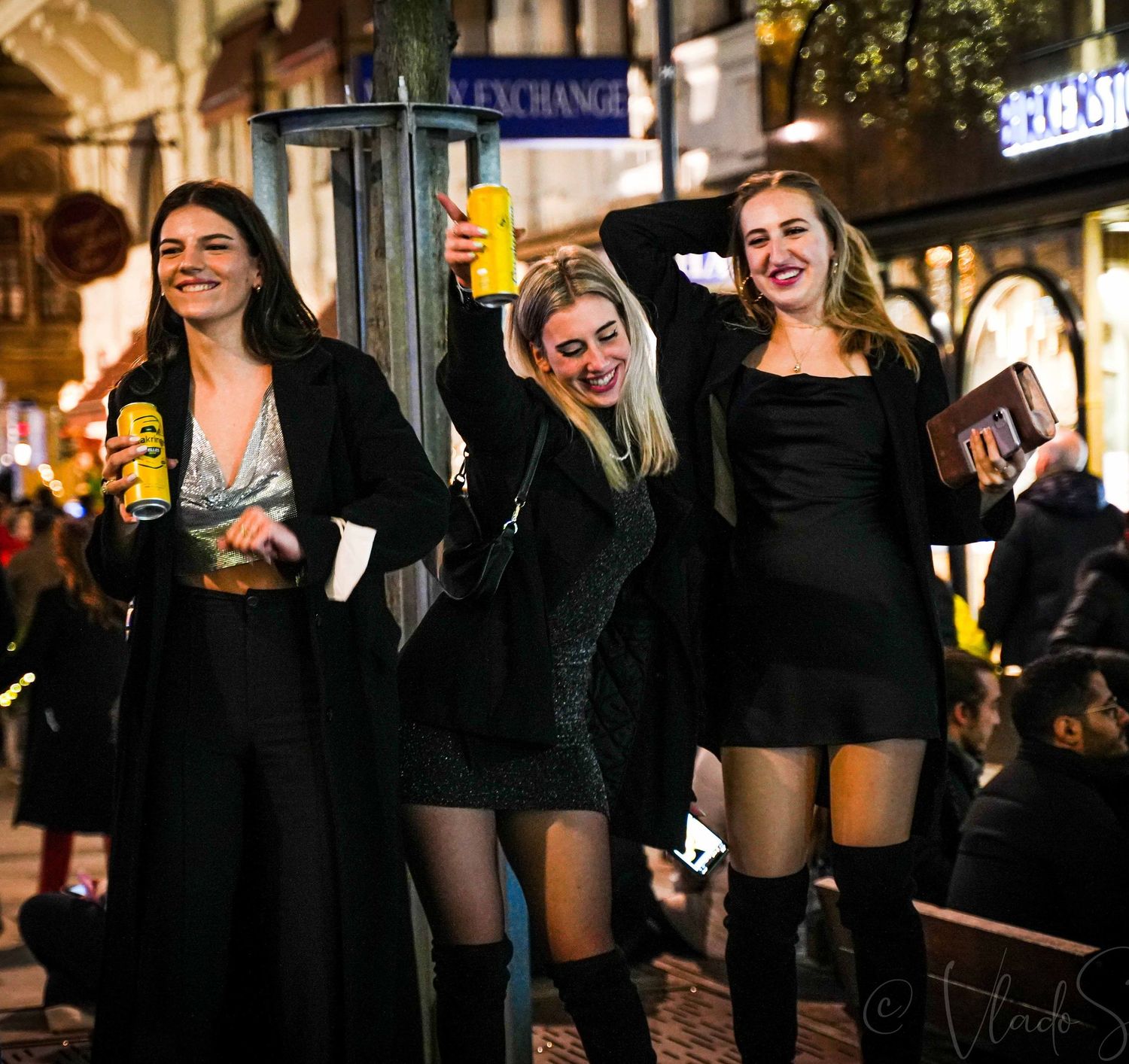 Three smiling women in black outfits celebrating on a lively night out on a busy city street.