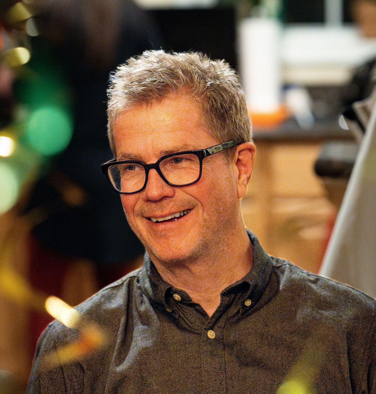 Middle-aged man with gray hair and black glasses smiling warmly in a social setting.