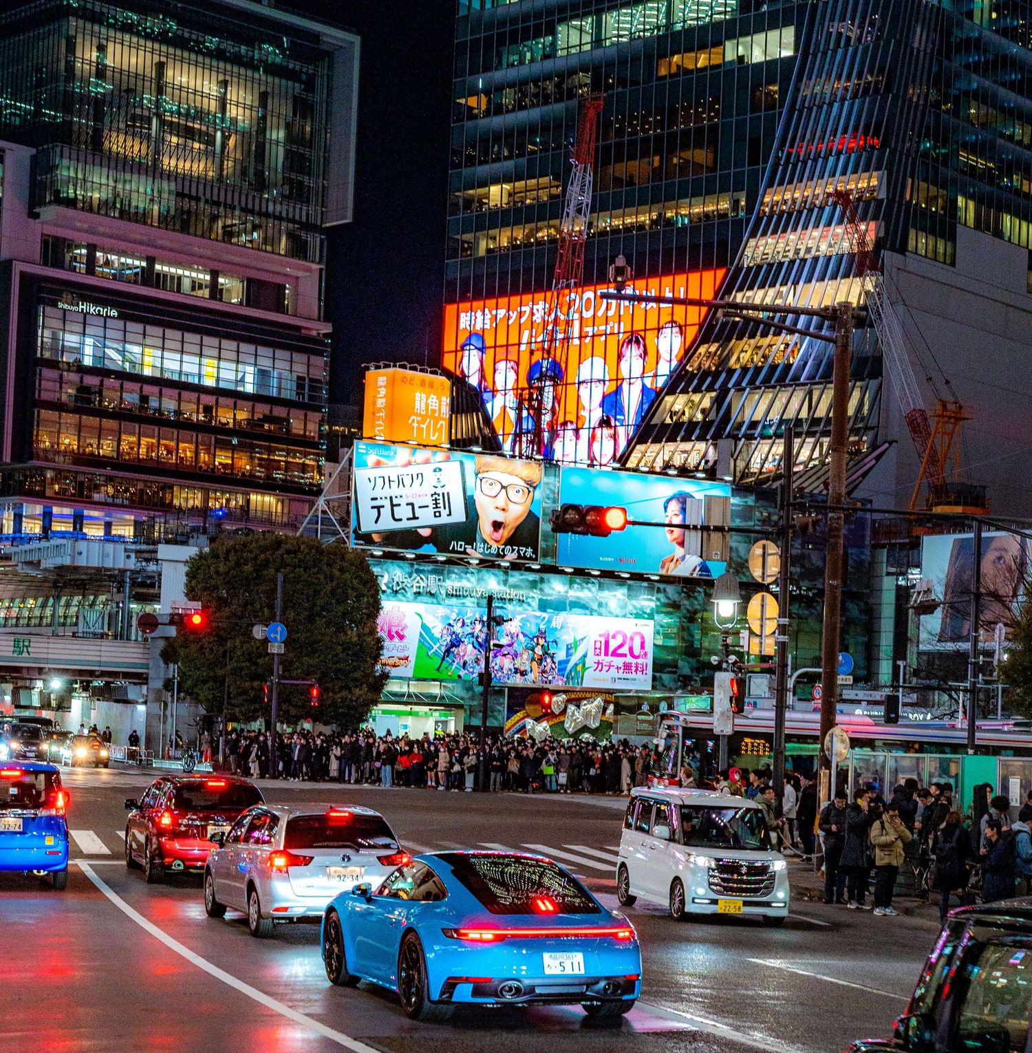 Busy Tokyo intersection at night with neon billboards, crowds, and a blue Porsche sports car in Shibuya district.