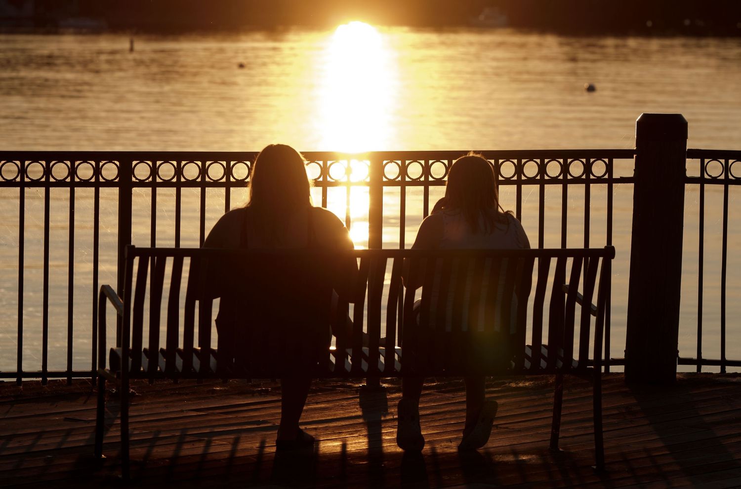 Two silhouetted figures sit on a bench watching a golden sunset over a calm waterfront behind an iron railing.