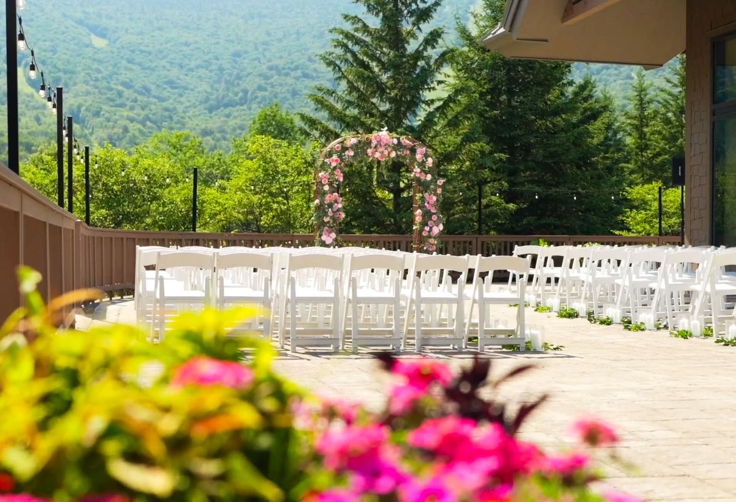 Outdoor wedding ceremony setup with white chairs, floral arch, and lush green mountain backdrop.