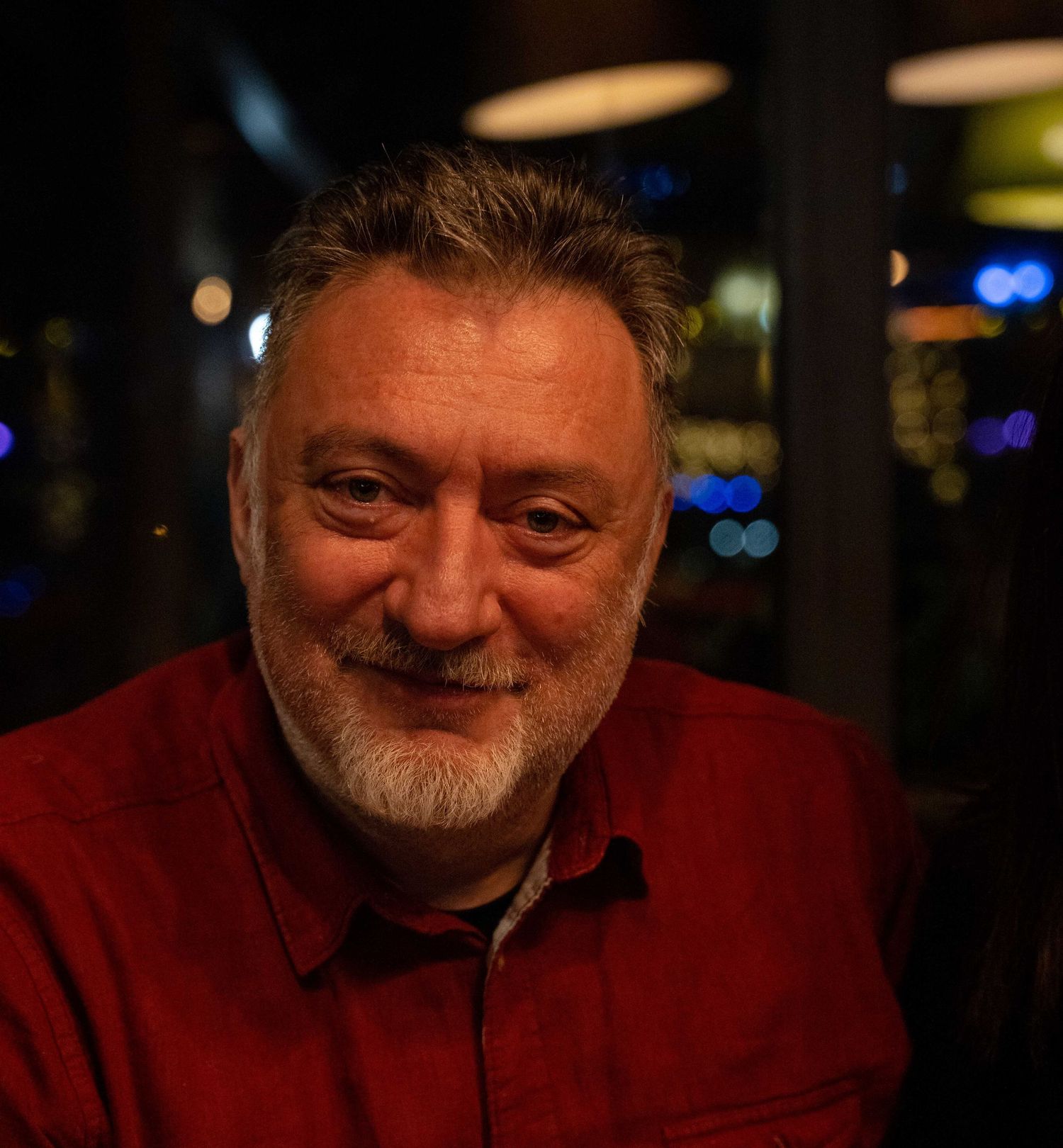 Smiling middle-aged man with gray beard wearing a red shirt in a restaurant at night.