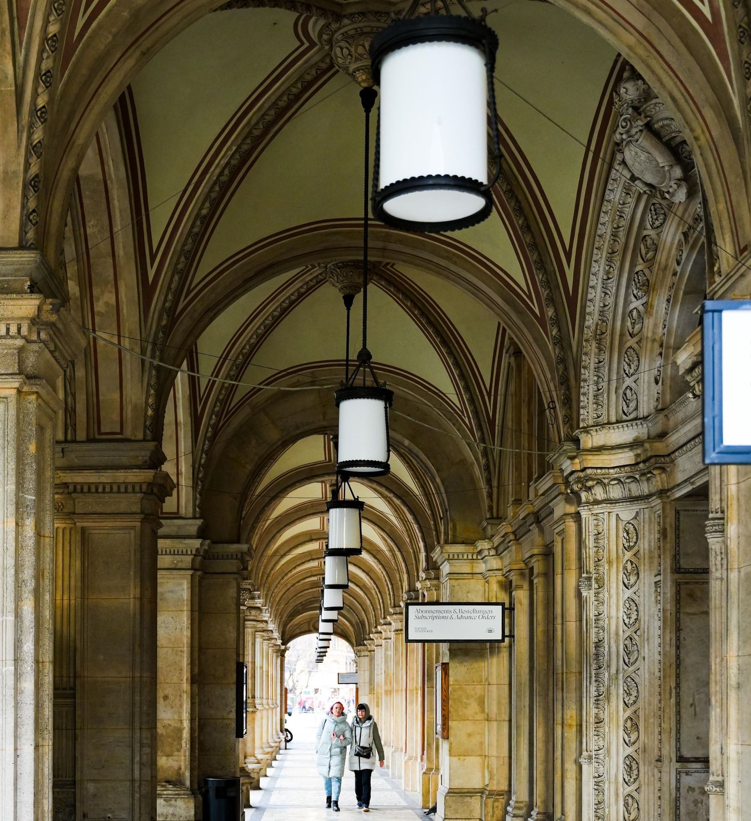 Grand arched corridor with ornate stone columns and hanging lanterns in a historic European building.