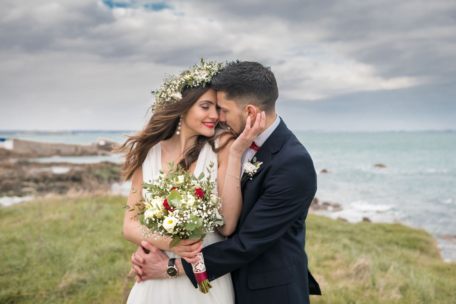 Romantic bride and groom embracing by the sea, bride holding wildflower bouquet with floral crown.
