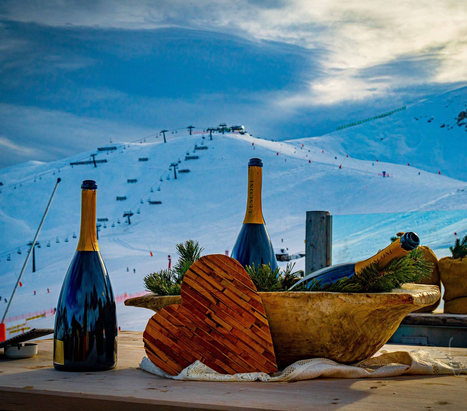 Champagne bottles in a wooden bowl on a snowy ski resort terrace with ski slopes and chairlifts in background.