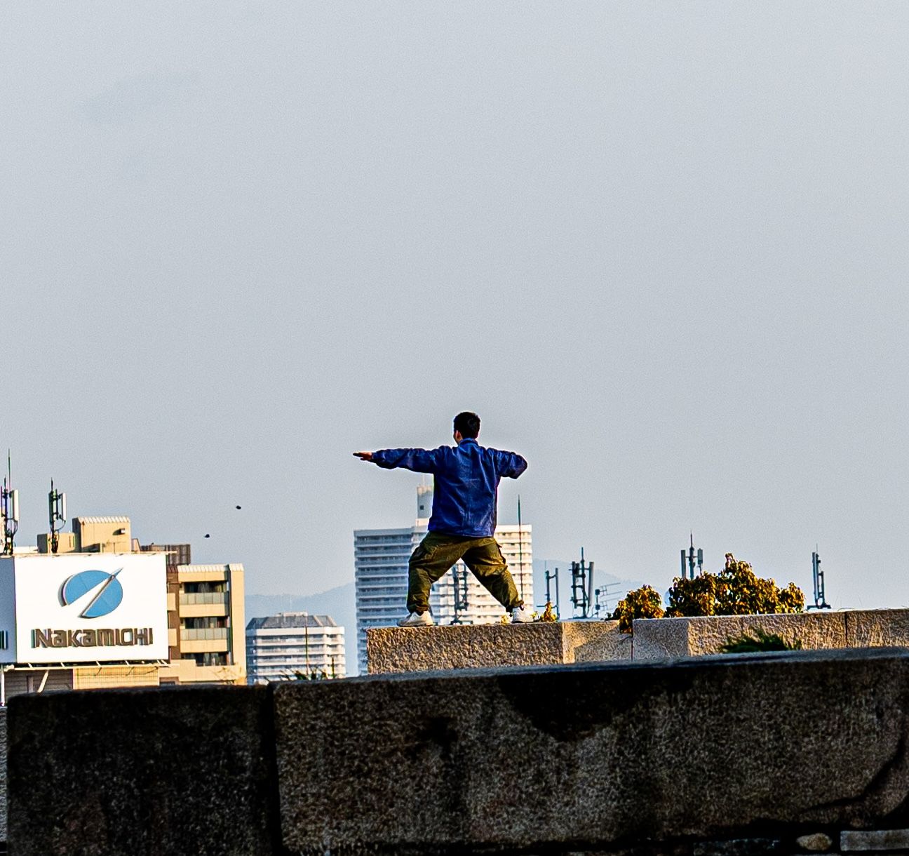 A man in a blue jacket stands with arms outstretched on a concrete wall against an urban city skyline.
