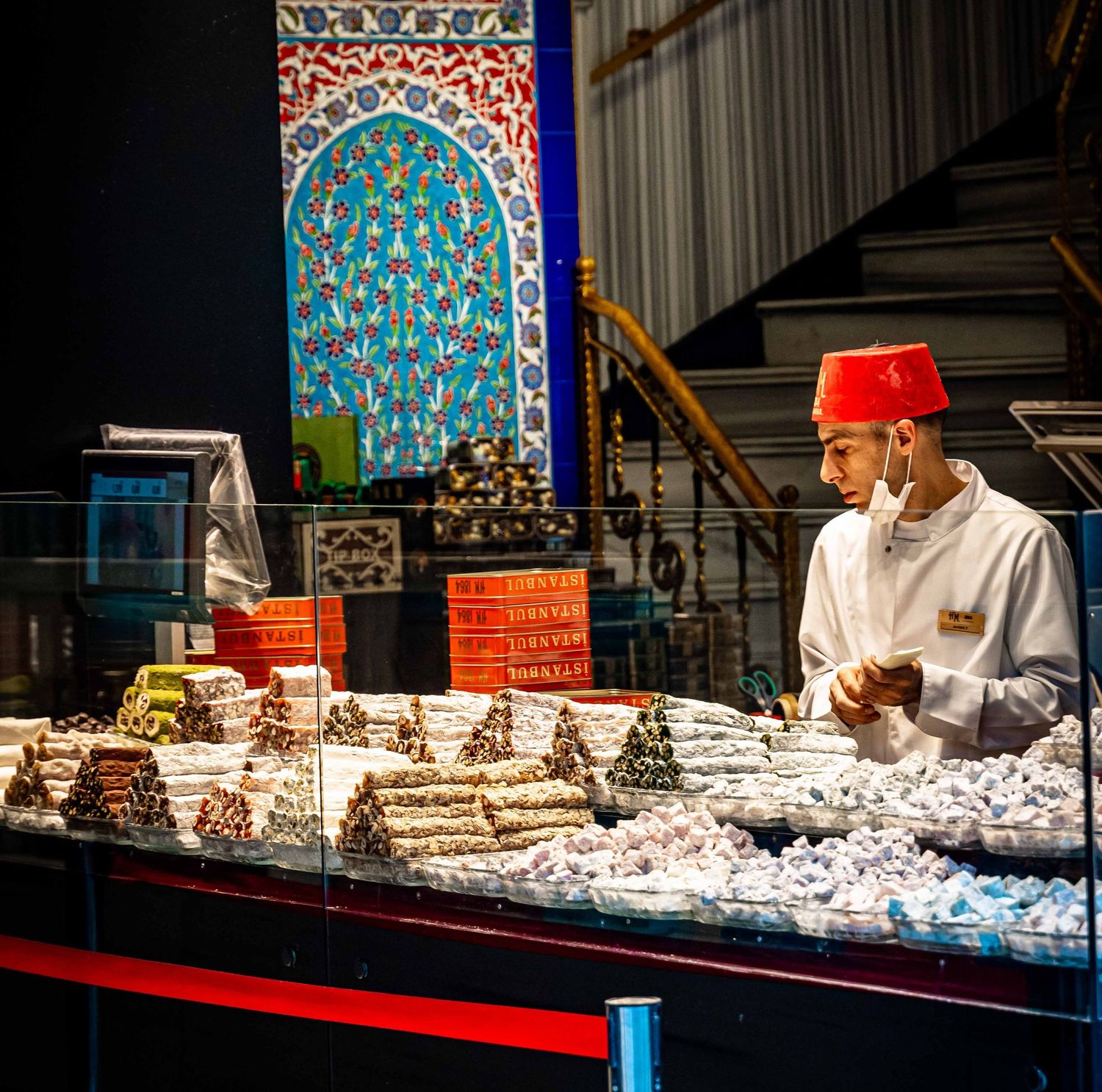 Vendor in a red fez hat selling colorful Turkish delight at a traditional market stall.