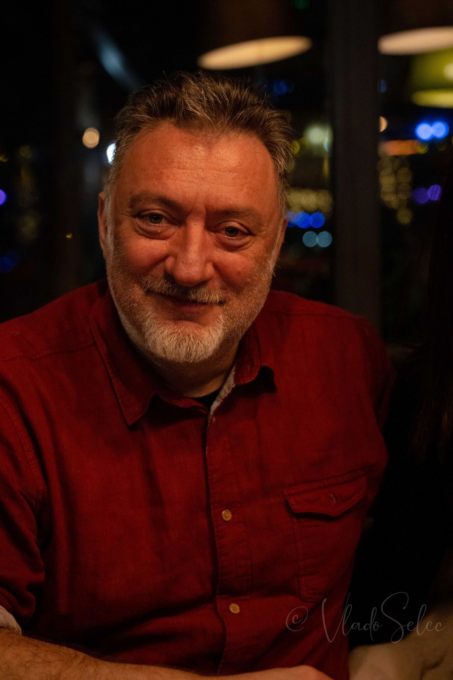 Smiling middle-aged man with gray hair and beard wearing a red shirt in a dimly lit restaurant.