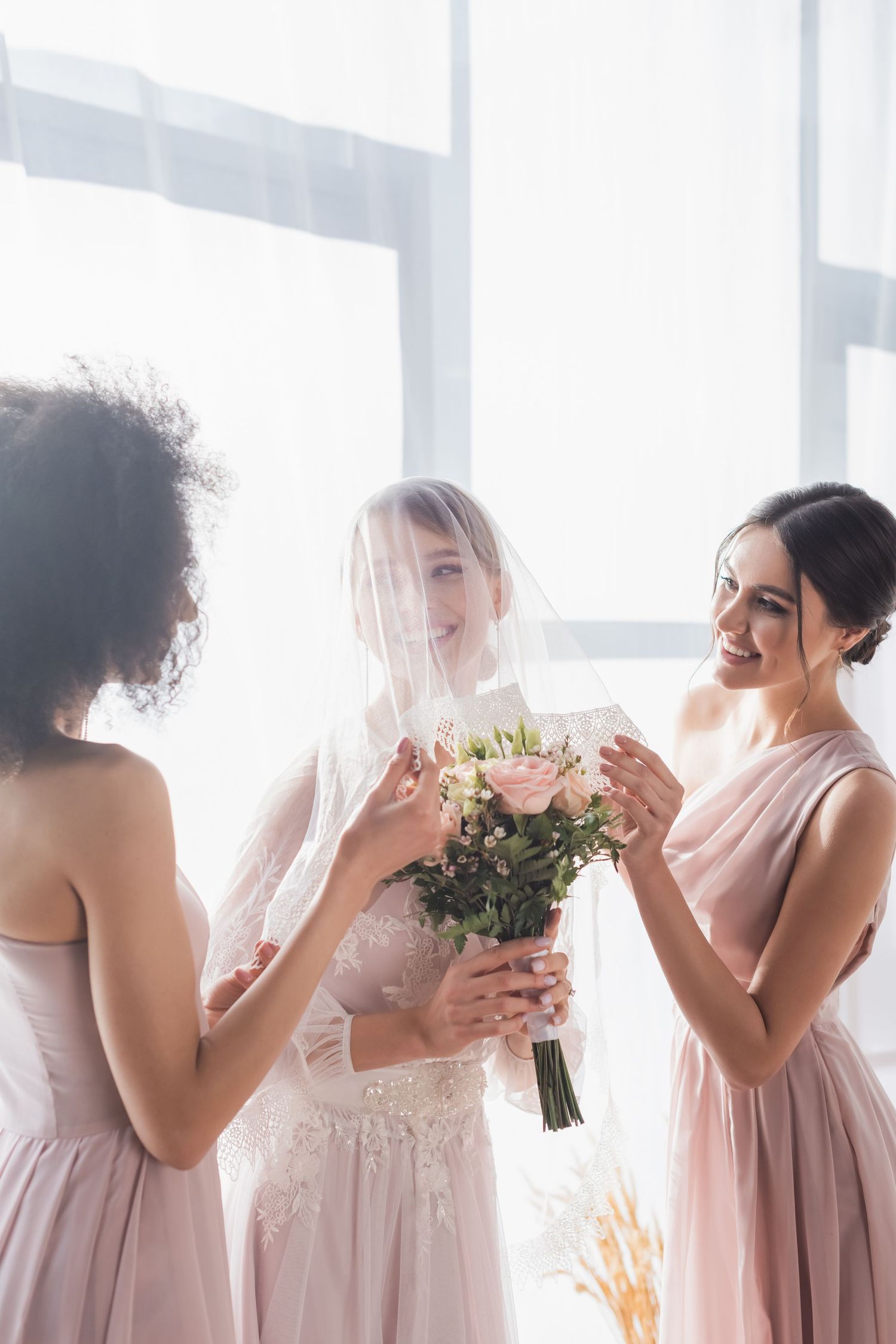 Bride in white lace gown with veil holding bouquet surrounded by smiling bridesmaids in pink dresses.