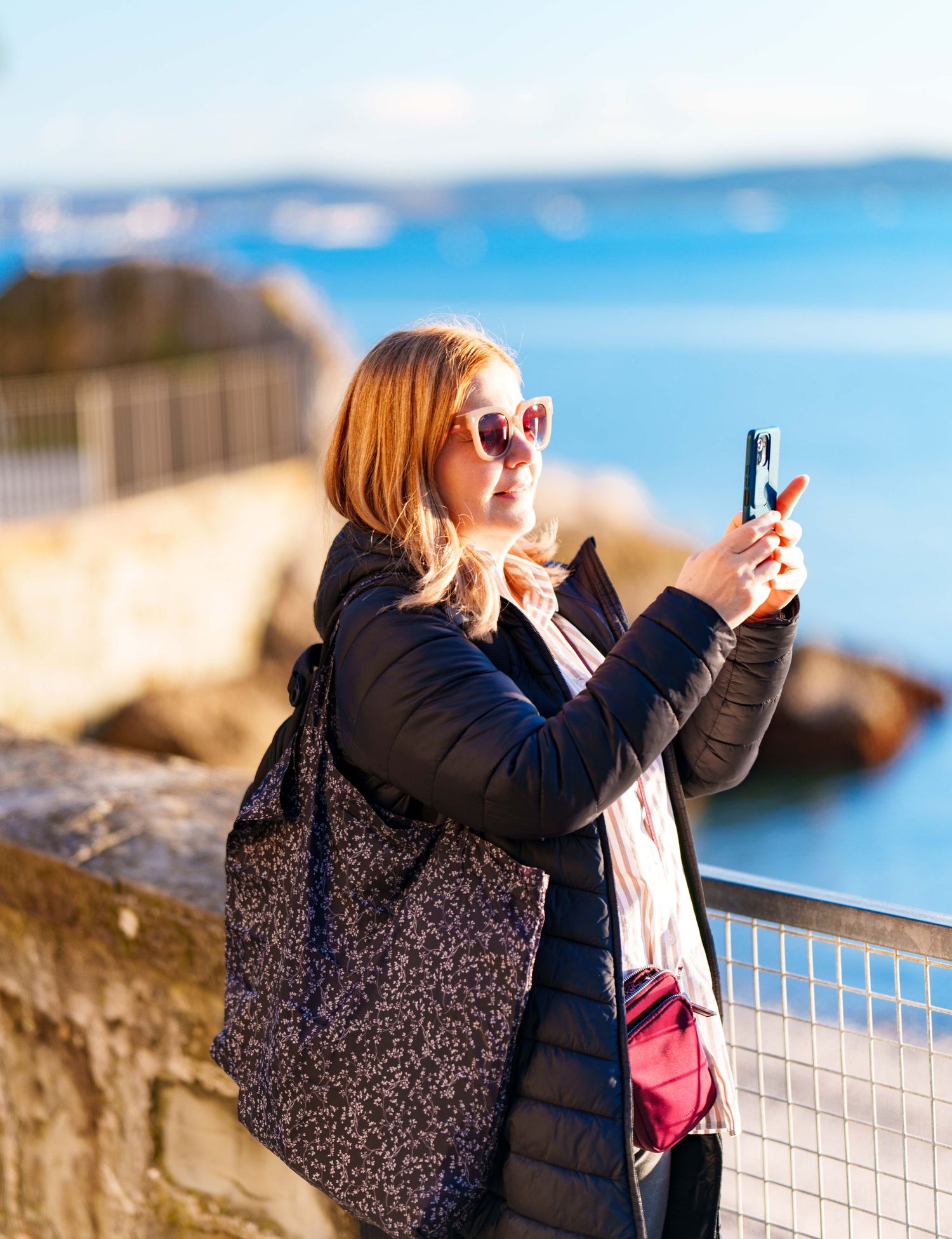 Blonde woman in sunglasses taking a smartphone photo by a coastal wall on a sunny day.
