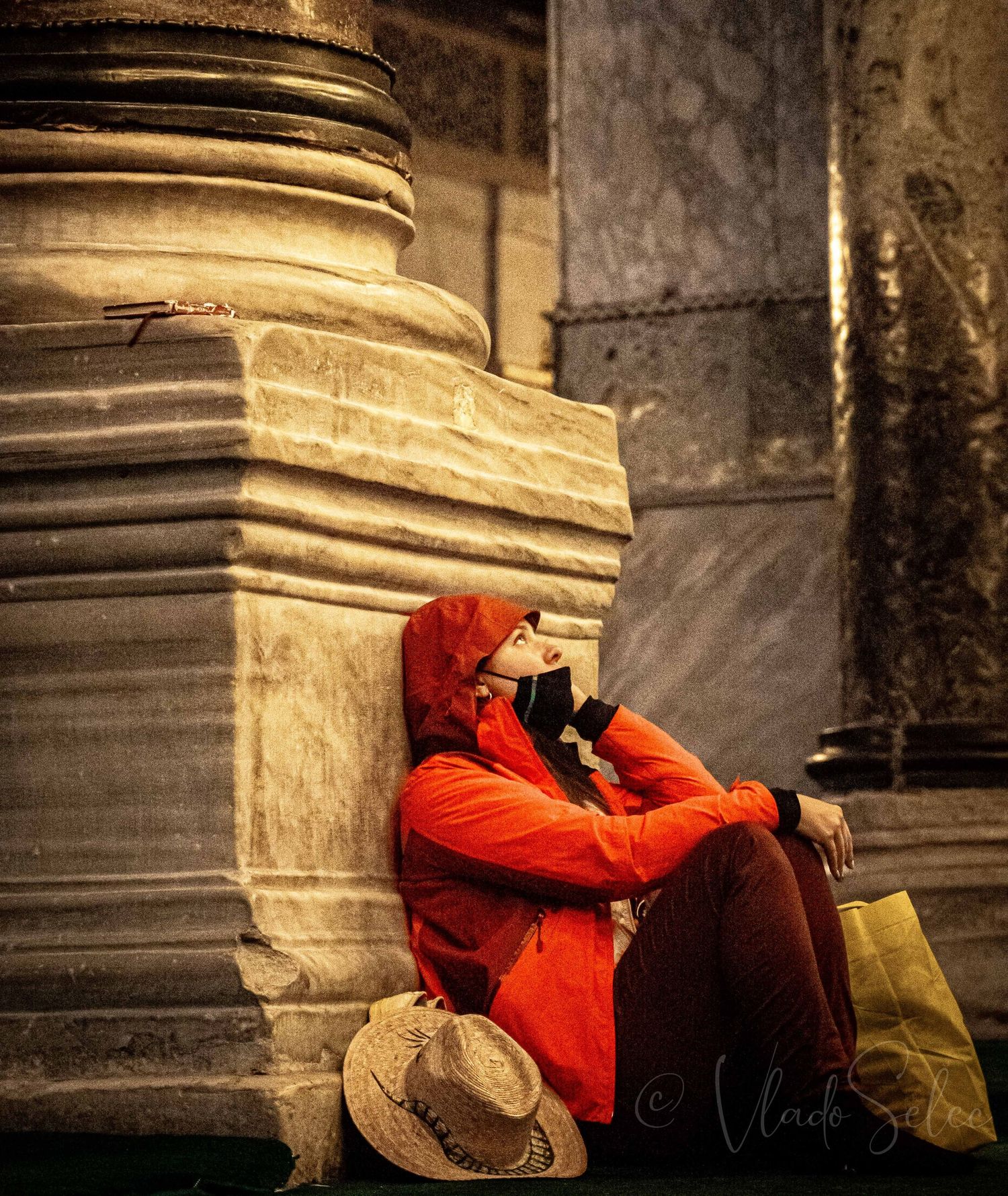 Person in red hooded jacket sitting against marble column wearing mask with hat and shopping bag.