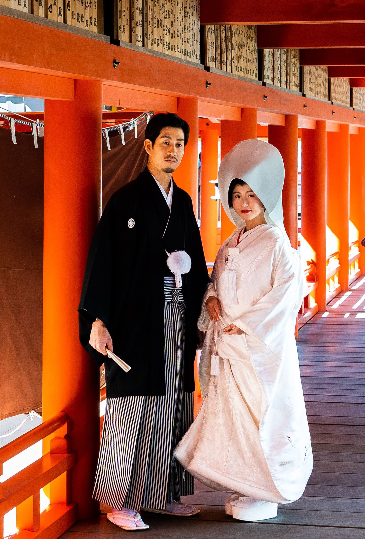 Japanese couple in traditional wedding attire posing in a red shrine corridor during a Shinto ceremony.
