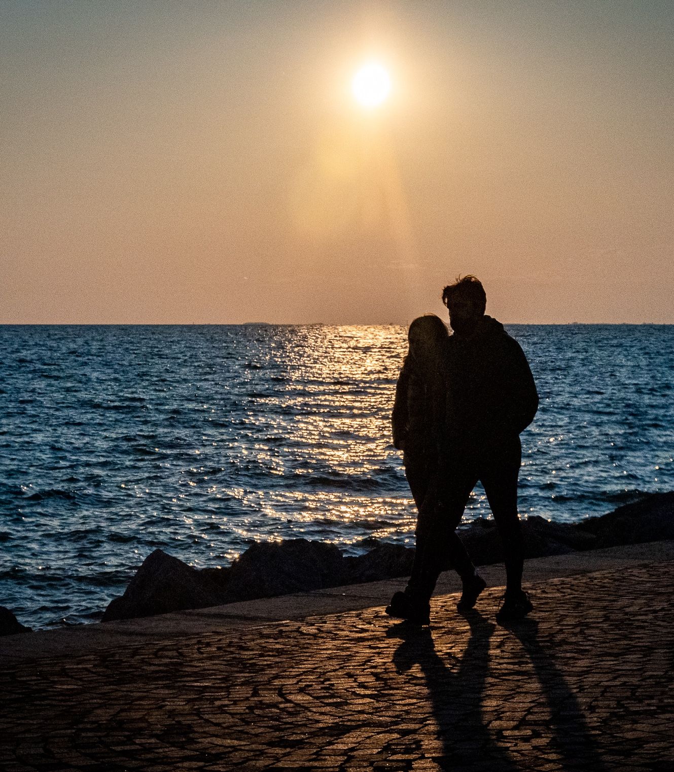 Romantic couple silhouetted against golden sunset over calm ocean waters on a cobblestone waterfront promenade.