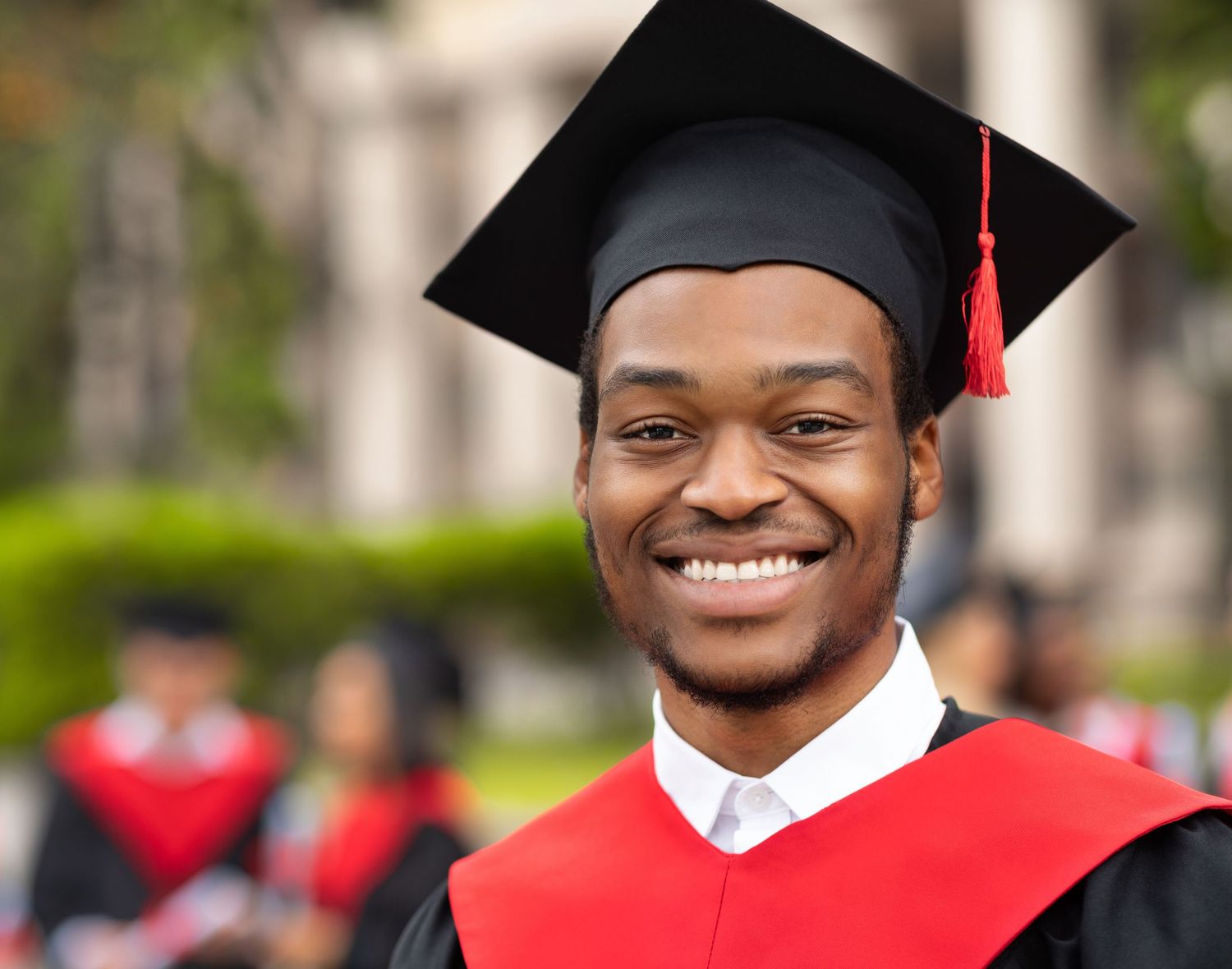 Happy young Black male graduate smiling in cap and gown with red accents outdoors.
