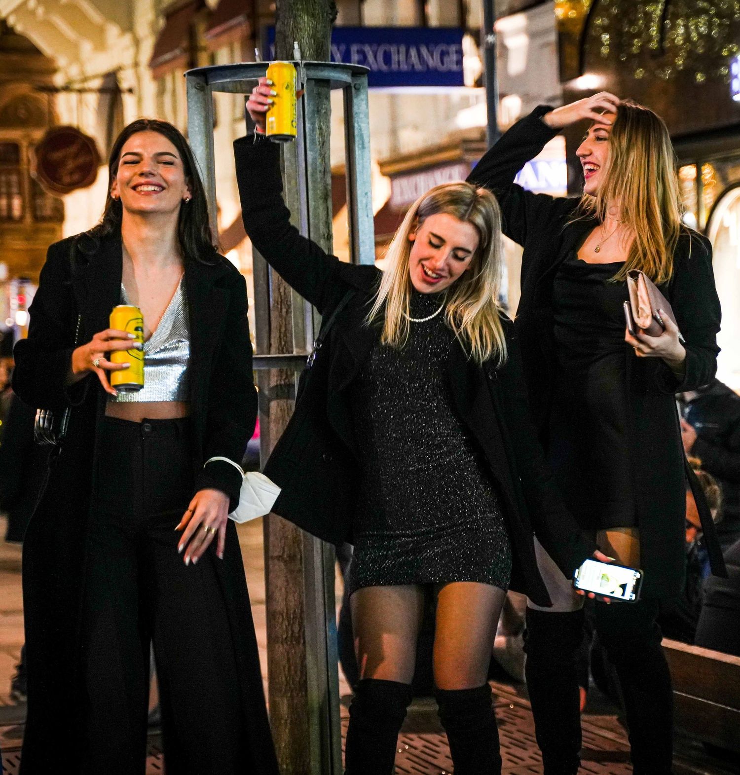 Three young women in black outfits celebrating outdoors at night holding drinks.