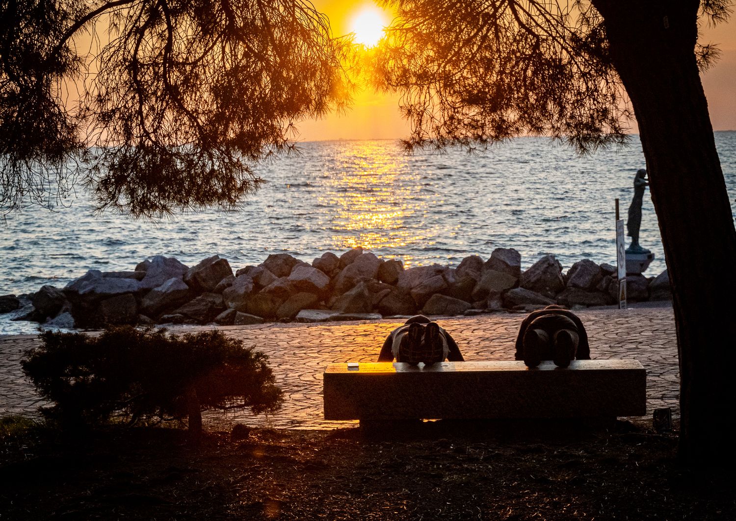 Two people sitting on a bench under pine trees watching a golden sunset over a rocky Mediterranean coastline.