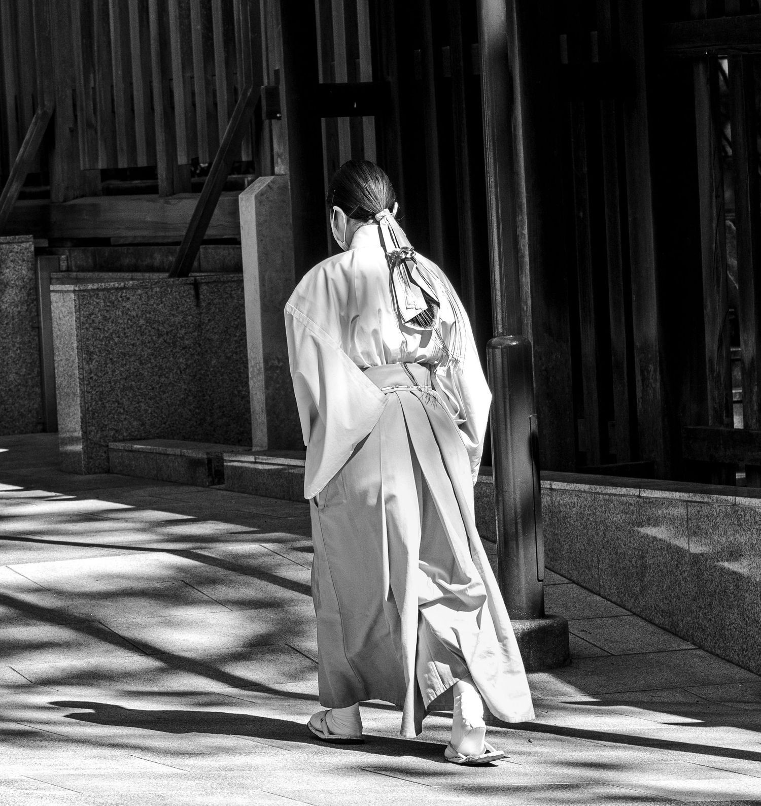 A shrine maiden in traditional white hakama walks away through a sunlit Japanese temple corridor in black and white.