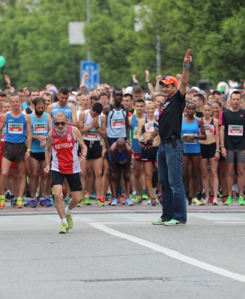 Runners at the start of a marathon race with a Serbia athlete and official signaling the beginning.