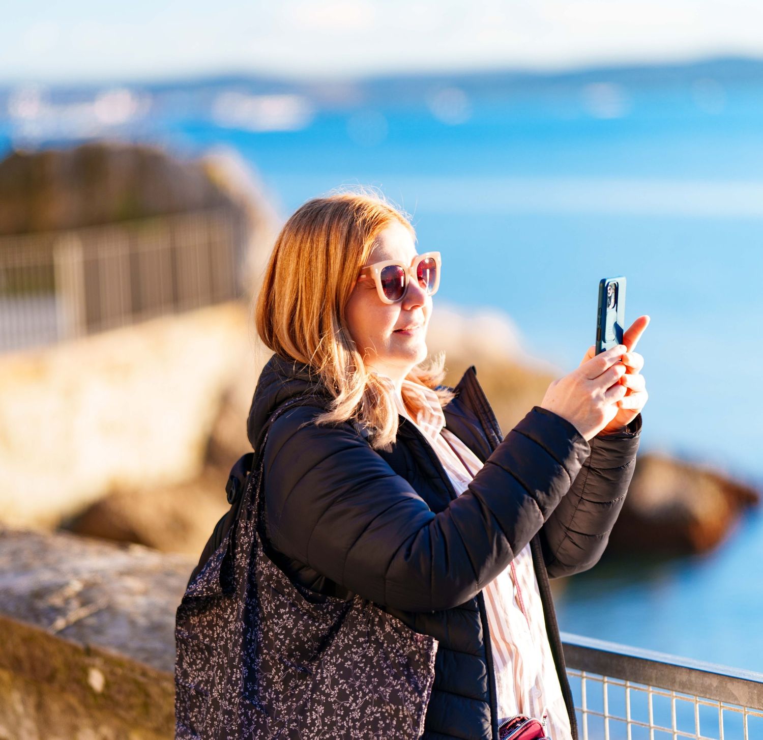 Blonde woman in sunglasses taking a photo with smartphone near a coastal waterfront.