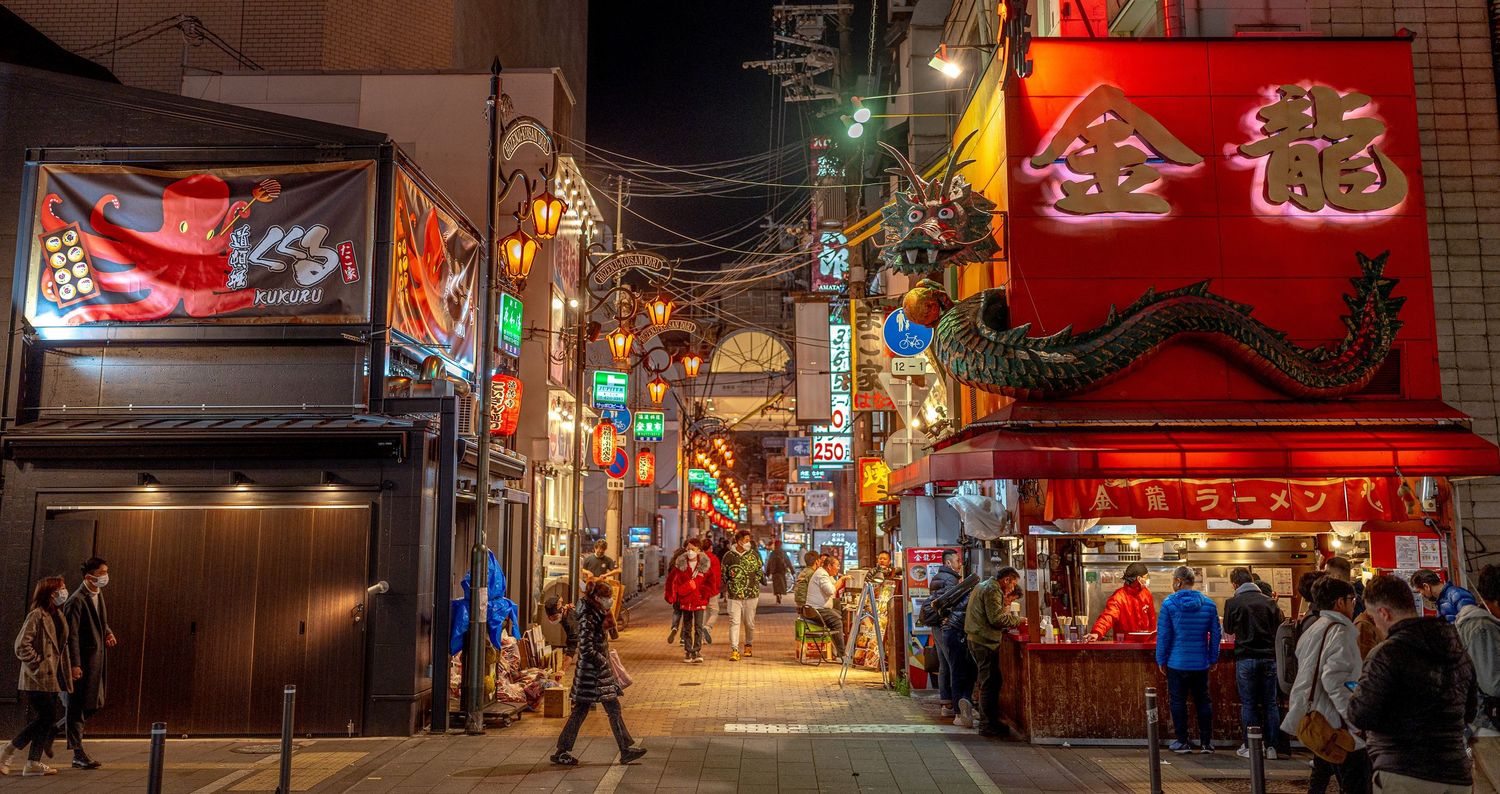 Busy Japanese Chinatown alley illuminated by colorful neon signs and lanterns at night.