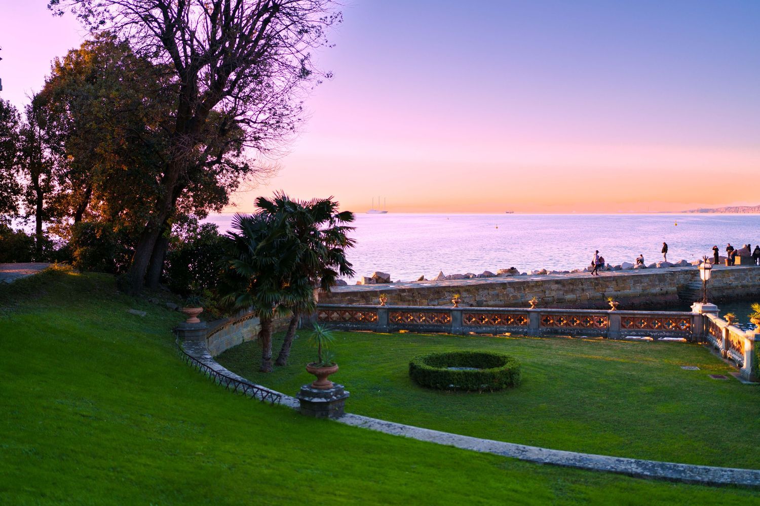 Lush garden terrace at sunset with manicured lawn and panoramic ocean views under a pink sky.