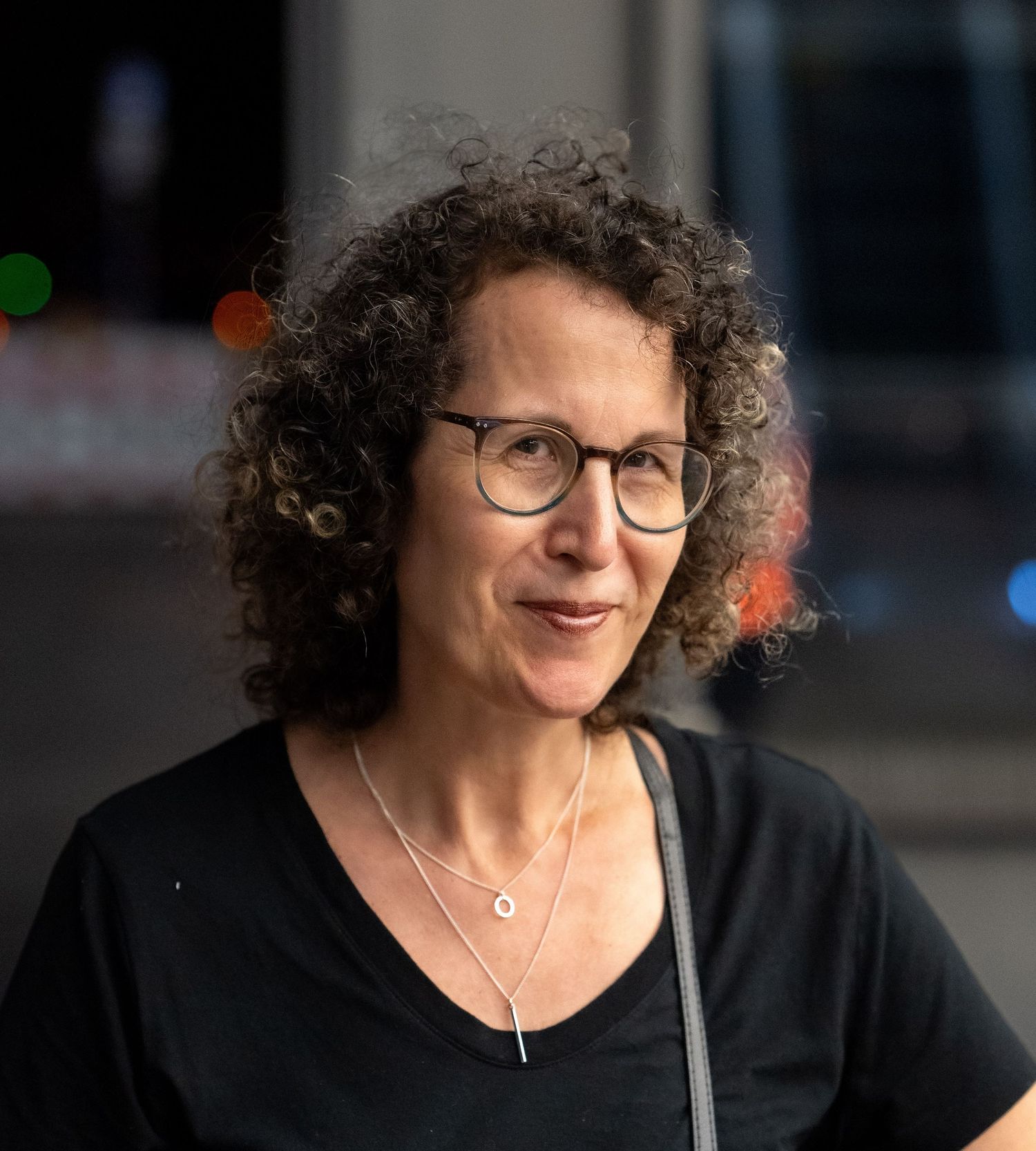 Middle-aged woman with curly hair and round glasses wearing black top outdoors at night.