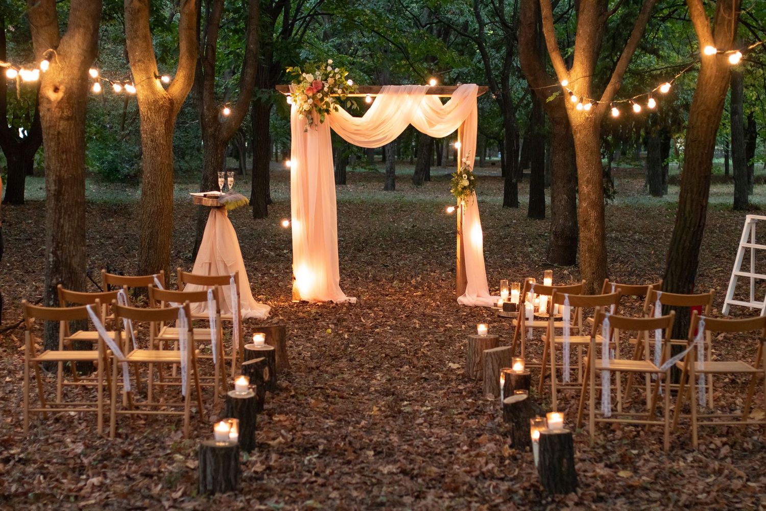 Enchanting outdoor wedding ceremony setup in a forest with candles, string lights, and a draped arch.