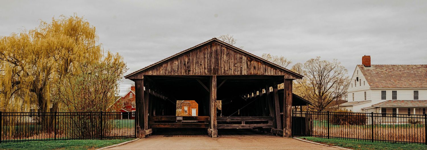 A rustic wooden covered bridge structure with an open passage framed by autumn trees and a white farmhouse.