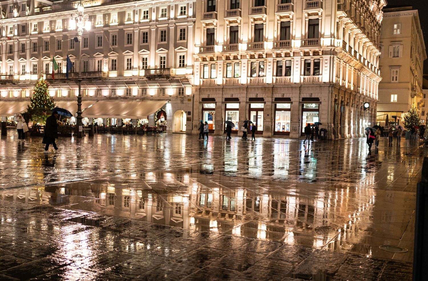 A rain-soaked European plaza glows with warm golden reflections from ornate illuminated buildings at night.