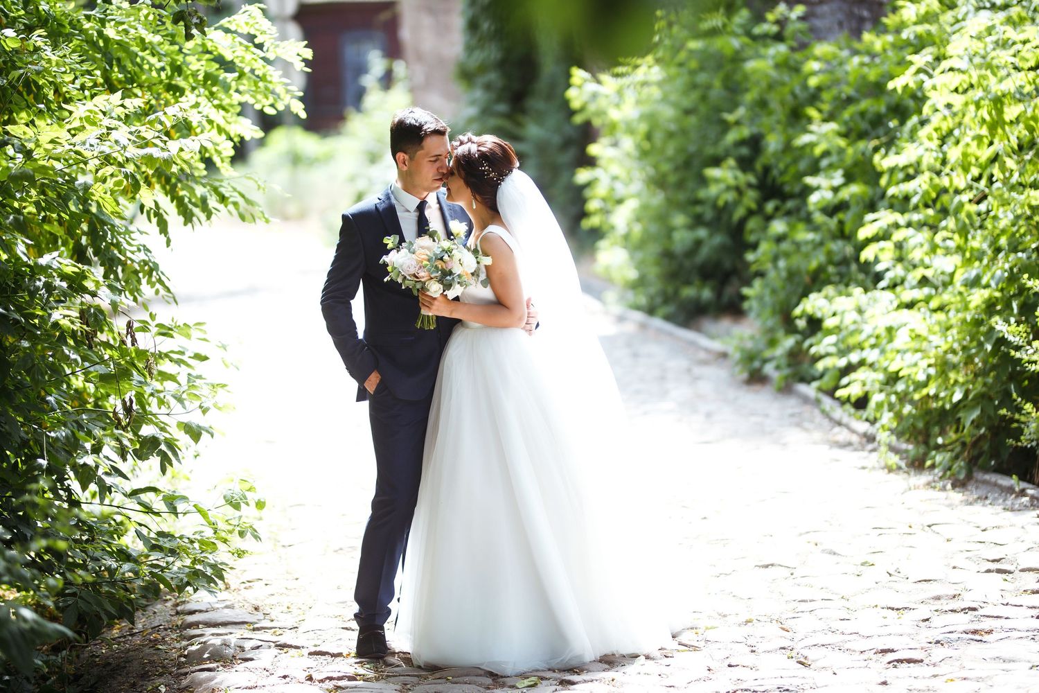 Newlywed couple sharing a romantic kiss on a sunlit garden path surrounded by lush green foliage.