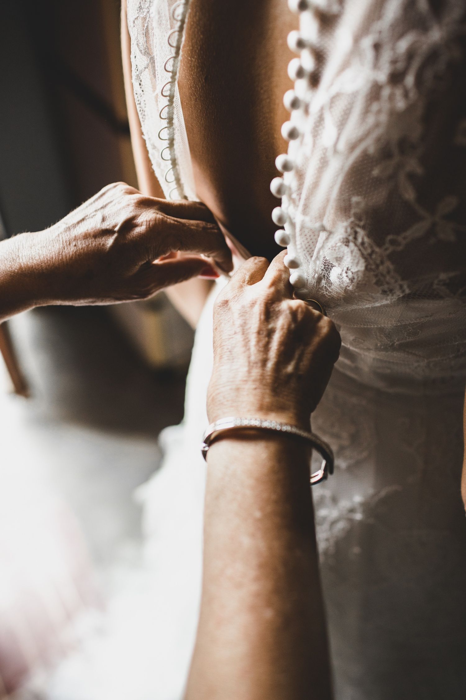 Hands fastening pearl buttons on a white lace wedding dress, close-up bridal preparation photo.