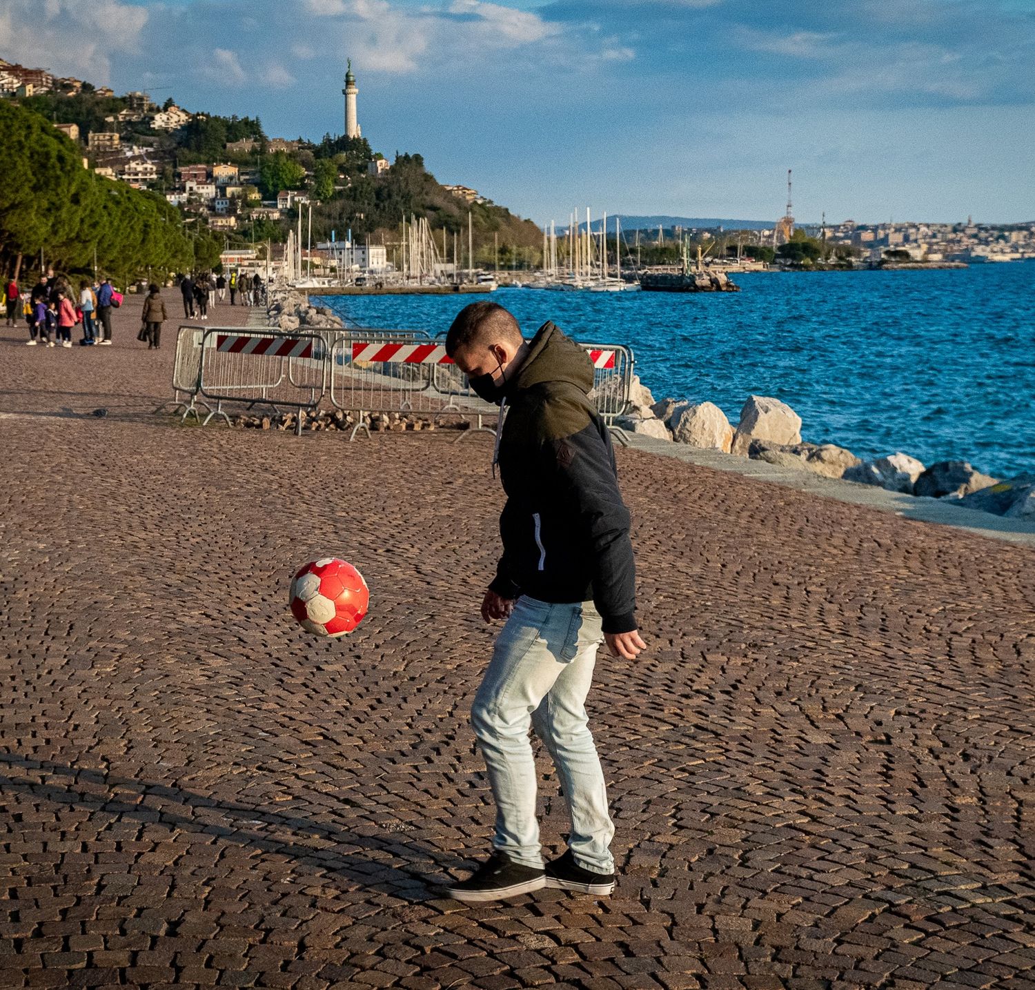 Young man juggling a soccer ball on a cobblestone waterfront promenade with harbor view.