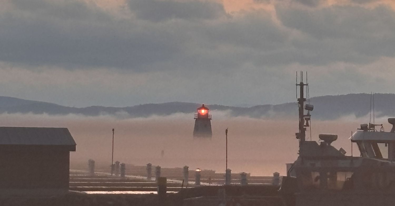 Misty harbor scene with a glowing red lighthouse beacon visible through morning fog.