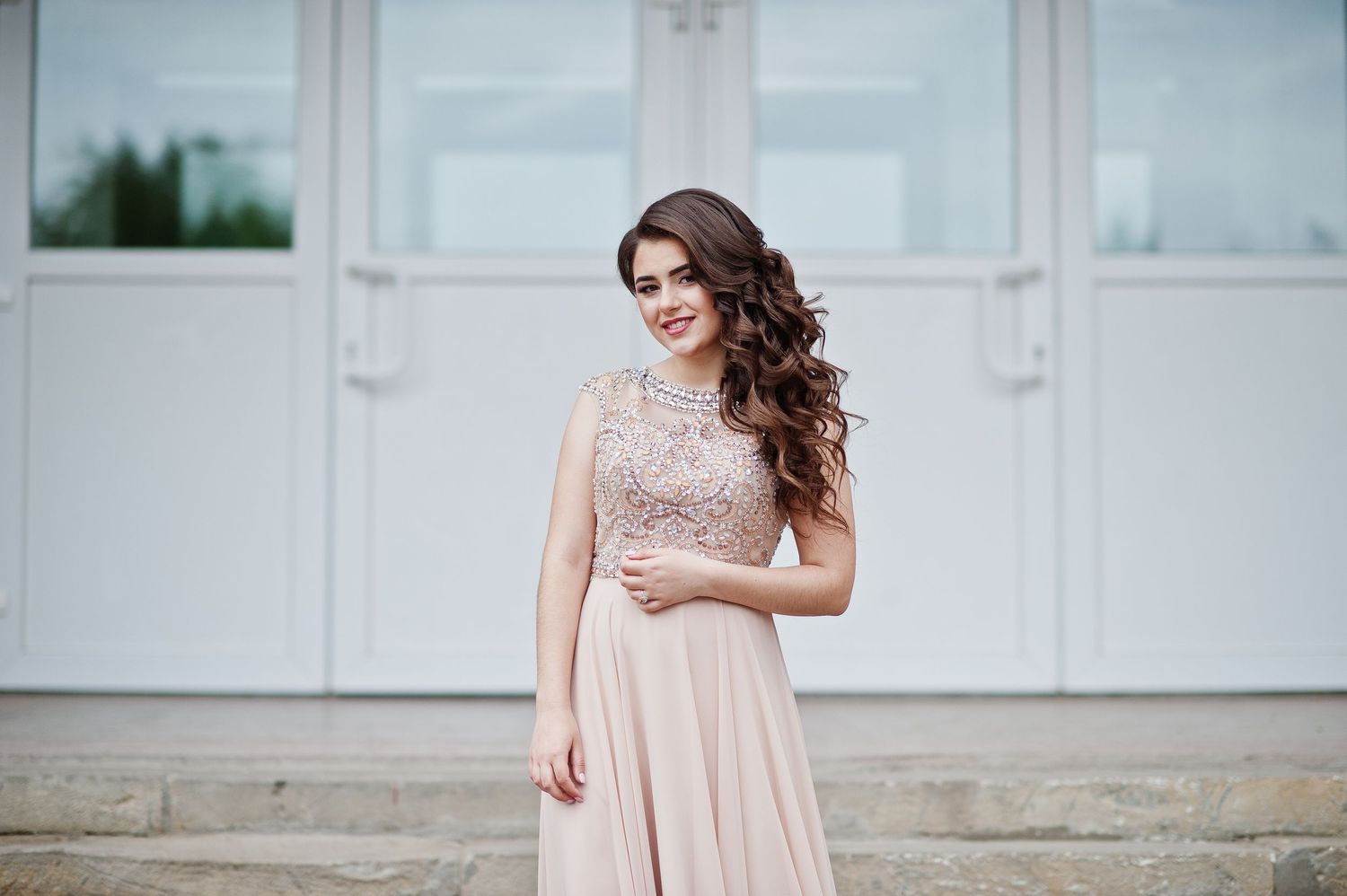 Beautiful young woman in an elegant blush pink evening gown posing outdoors near white doors.