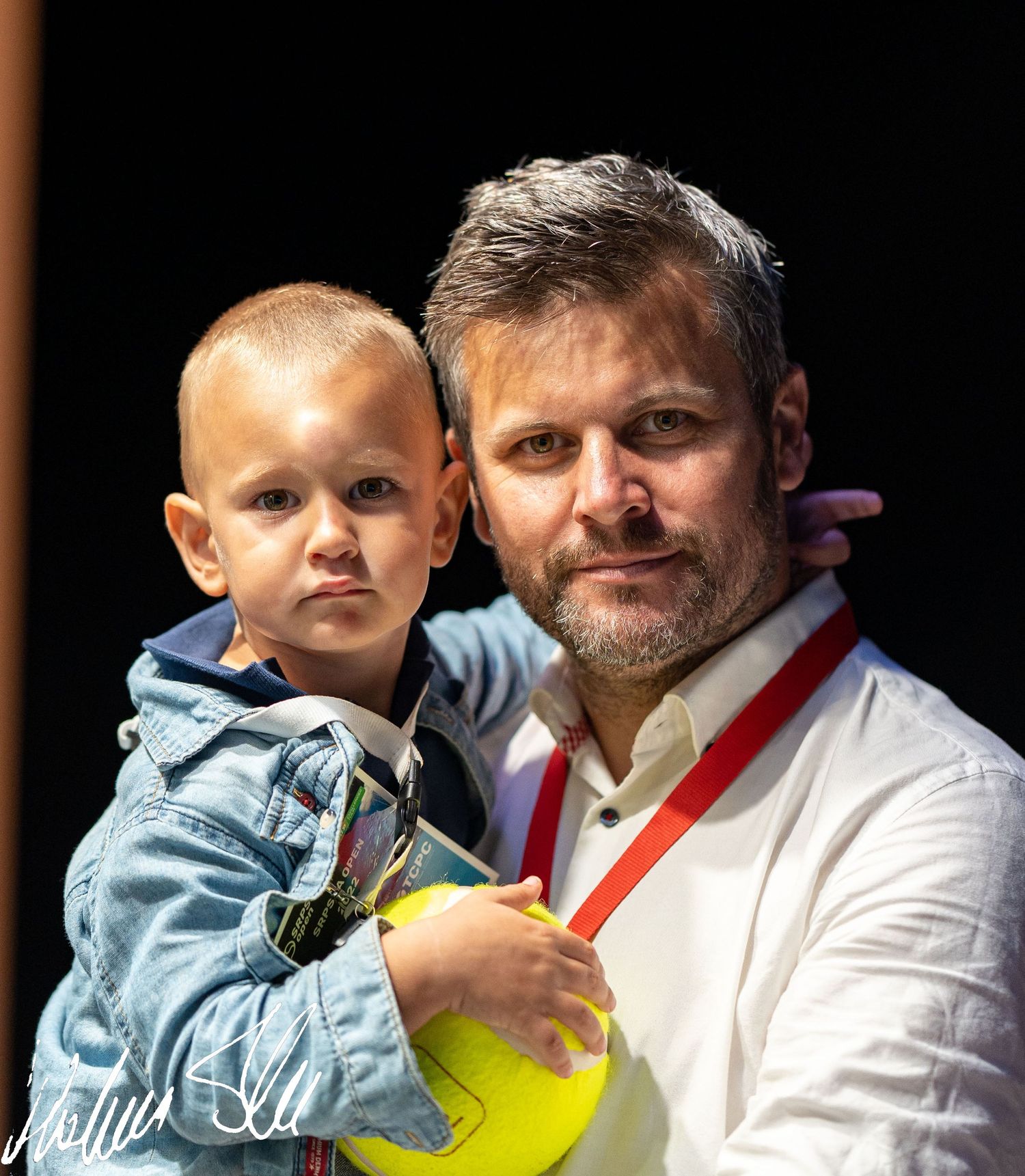 Man with beard holding a young child with a yellow ball at an indoor event.