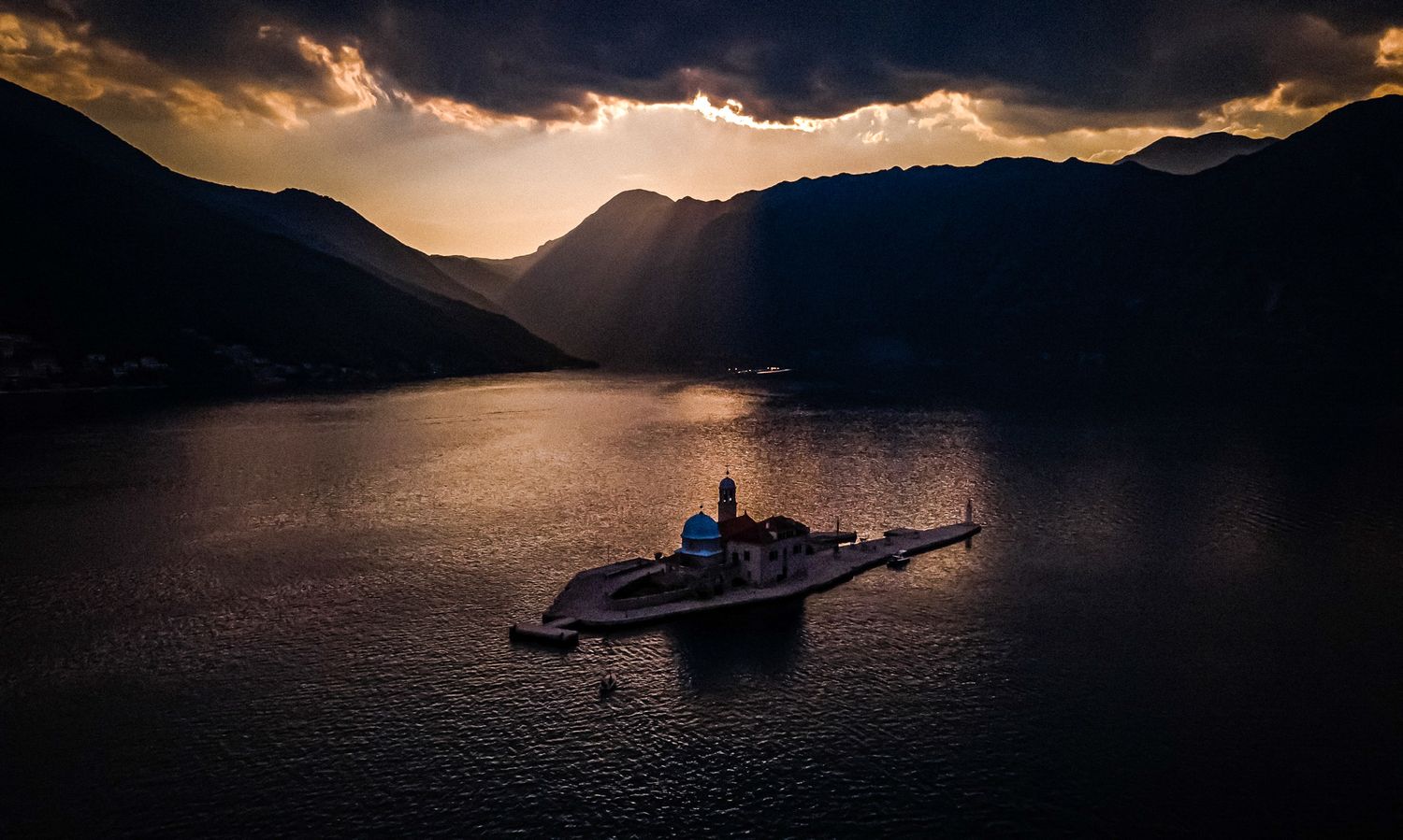 Aerial view of a small island monastery on a calm bay with dramatic sunbeams and dark mountain backdrop.