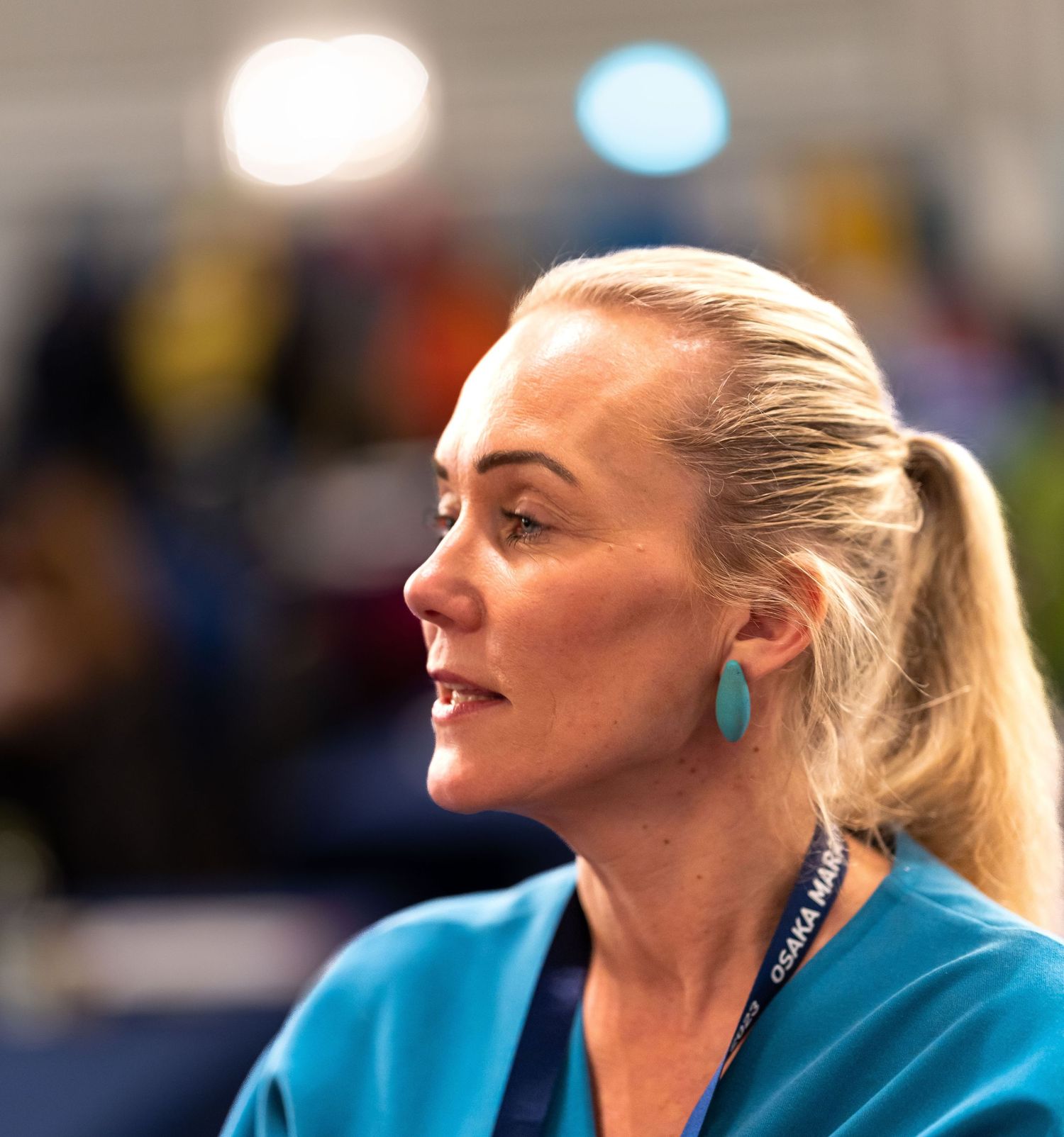A blonde woman in teal scrubs wearing turquoise earrings and a lanyard looks sideways in a busy indoor setting.