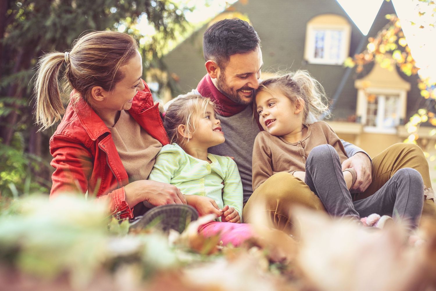 Happy family with two young daughters sitting together in autumn garden outside their home.
