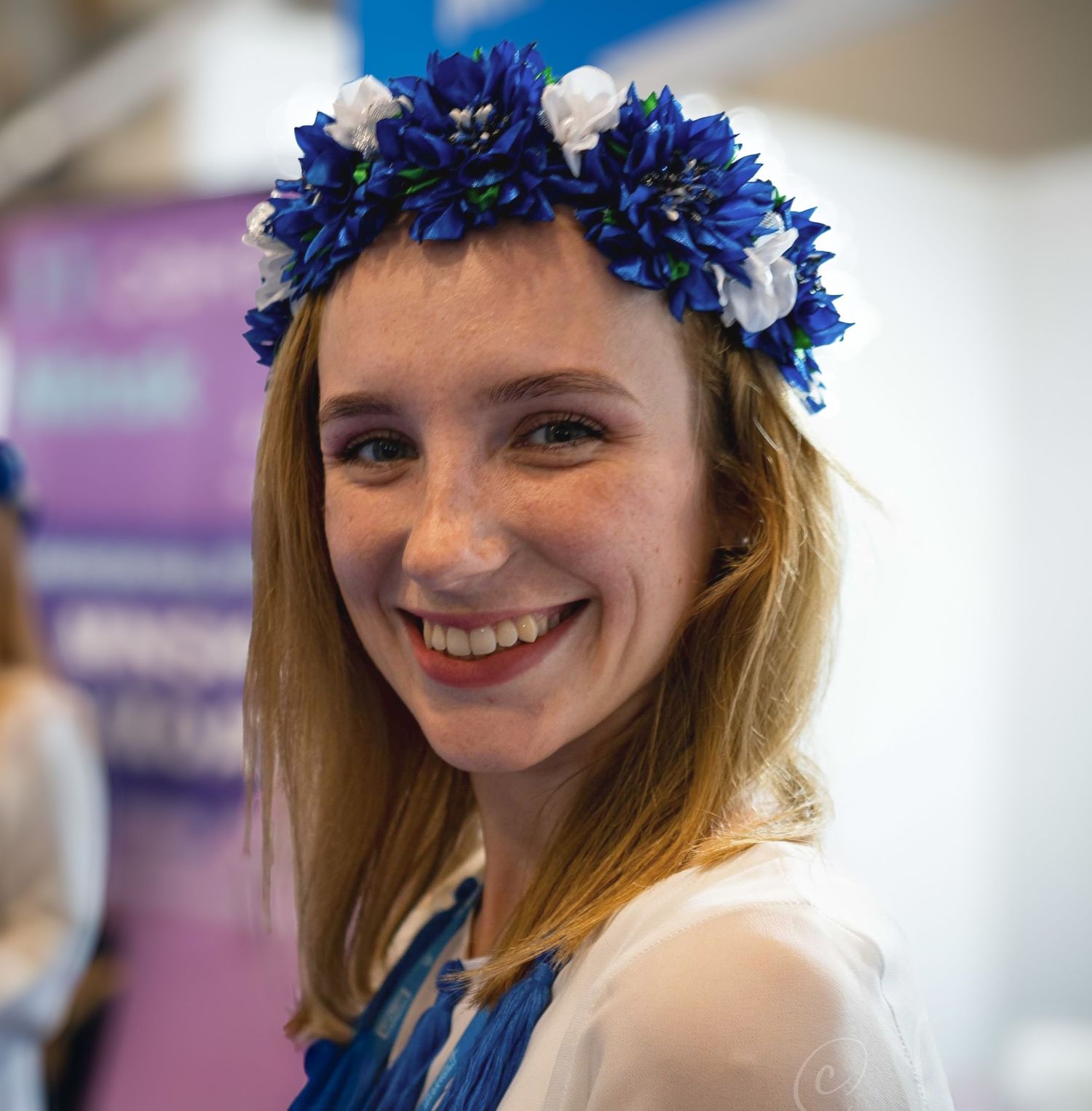 Smiling young woman wearing a blue and white floral crown at an indoor event.