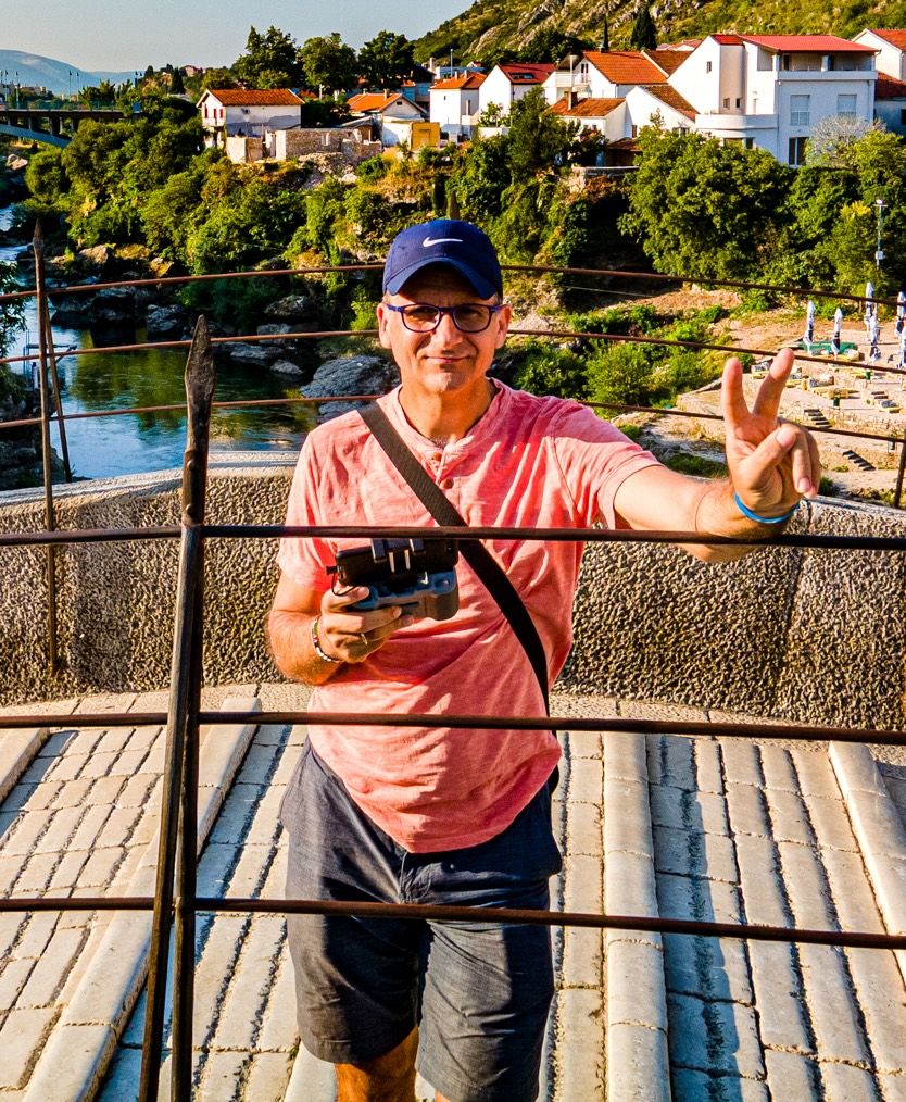 Man in pink shirt and blue cap holds camera, flashing peace sign on bridge overlooking coastal village.