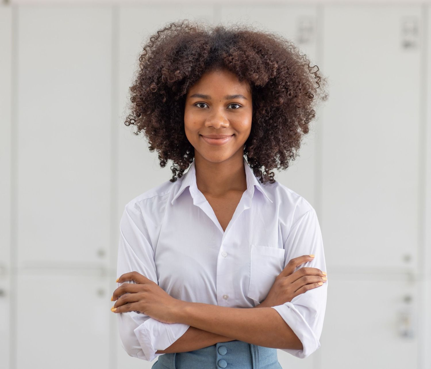 Confident young Black woman with curly hair standing with arms crossed in a white shirt.